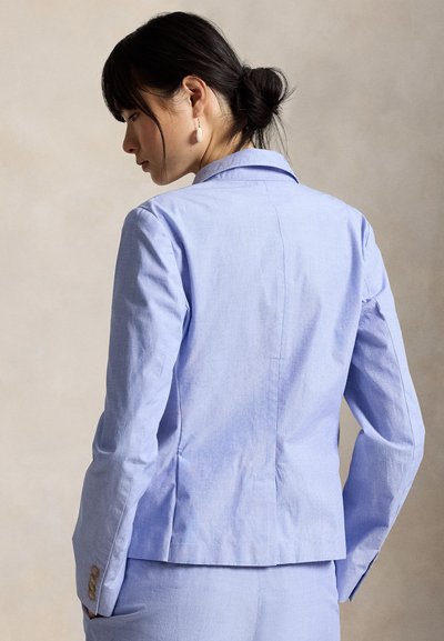 Γυναίκα με σκούρα μαλλιά συλλεγμένα σε κότσο φοράει ένα κομψό γαλάζιο σακάκι και παντελόνι που ταιριάζουν, στραμμένη με την πλάτη σε απλό μπεζ φόντο.