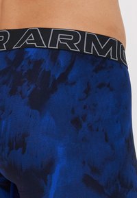 Plave sportske tajice sa uzorkom koje imaju crni elastični pojas s natpisom "ARMOUR" u sivoj boji. Glatka tekstura s uskim dizajnom.