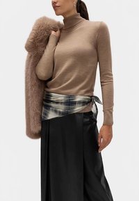 Pull à col roulé beige associé à un manteau en fausse fourrure. Écharpe à carreaux bleu marine et beige attachée à la taille, jupe noire élégante avec des plis.