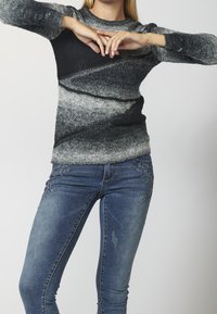 Femme portant un pull en maille dégradé noir et gris et un jean bleu brodé, les mains jointes au-dessus de la poitrine, sur un fond uni.