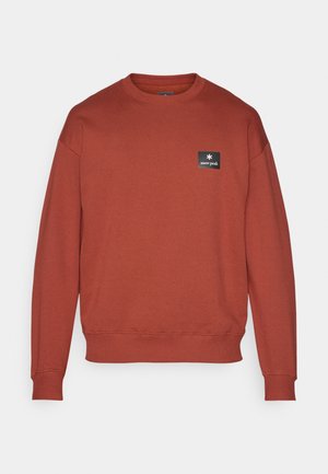 Sweatshirt couleur rouille en matériau doux, avec un col rond, des manches longues et un petit patch logo noir sur la poitrine.
