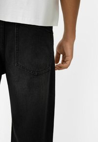 Los pantalones vaqueros de mezclilla negra tienen un corte casual con un detalle en el bolsillo trasero. Una camiseta blanca es parcialmente visible sobre los pantalones.