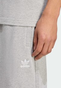 Šviesiai pilki medvilniniai sportiniai kelnės su šoninėmis kišenėmis, ant šlaunies puikuojasi baltas Adidas logotipas. Medžiaga atrodo minkšta ir tekstūruota.