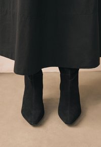 Schwarze Wildleder-Stiefeletten mit spitzer Zehenpartie, strukturierter Oberfläche und figurbetontem Design, teilweise verdeckt von einem schwarzen Kleid.