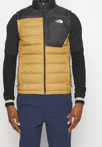 Gul och svart isolerad väst med vertikal quiltning, dragkedjeficka på bröstet och en logotyp på övre vänstra sidan. Buren över en svart långärmad topp.