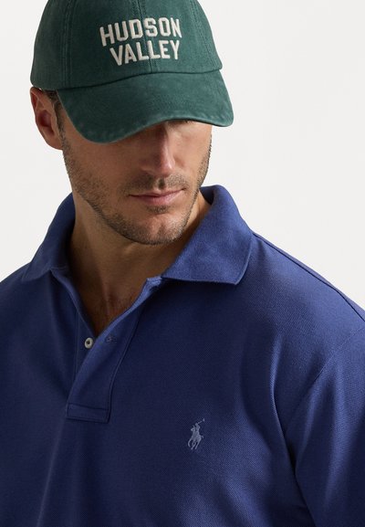 Μπλε πόλο μπλουζάκι με δύο κουμπιά και ένα μικρό λογότυπο, συνδυασμένο με πράσινο καπέλο που διαθέτει άσπρο ραμμένο κείμενο "HUDSON VALLEY."