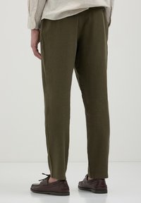 Pantalons en lin vert olive à coupe décontractée, avec un bouton à la taille, associés à des chaussures en cuir marron. Texture lisse, forme effilée.