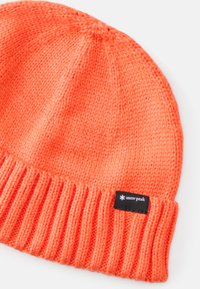 Knit beanie in bright orange, featuring a ribbed cuff and black branding tag with white logo. Soft, textured yarn throughout.