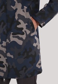 Maskovací bunda s tmavě modrými, černými a šedými vzory, se zipem a bočními kapsami, vyrobená z hladké, pružné látky.