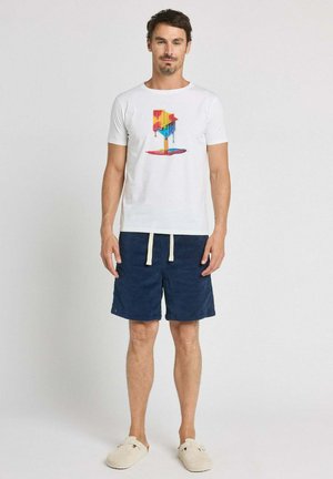 Homme portant un t-shirt blanc avec un graphisme coloré d'une boîte aux lettres fondante, un short bleu marine avec des cordons blancs et des chaussures beige à enfiler, debout.