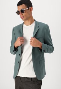 Tirkizna blazer s dva gumba, s izrezanim reverima i lijevom prsnom džepom. Nosi se preko svijetlosivog majice, uparen s crnim sunčanim naočalama.