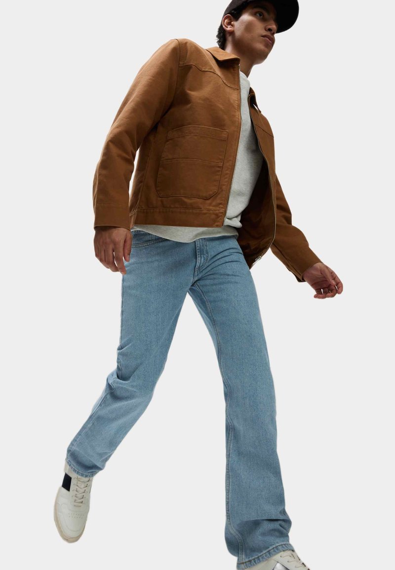 Chaqueta de algodón marrón con cierre de cremallera y bolsillos, llevada sobre un suéter gris y jeans azul claro, combinada con zapatillas blancas.