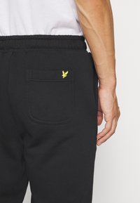 Czarne dresy wykonane z miękkiego materiału, z elastycznym pasem w talii, posiadające tylnią kieszeń ozdobioną małym żółtym logo.