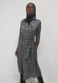 Schwarzes Kleid mit Punkten, langen Ärmeln, Knopfleiste und gebundenem Taillengürtel; aus einem glatten Stoff mit einer leicht fließenden Silhouette.