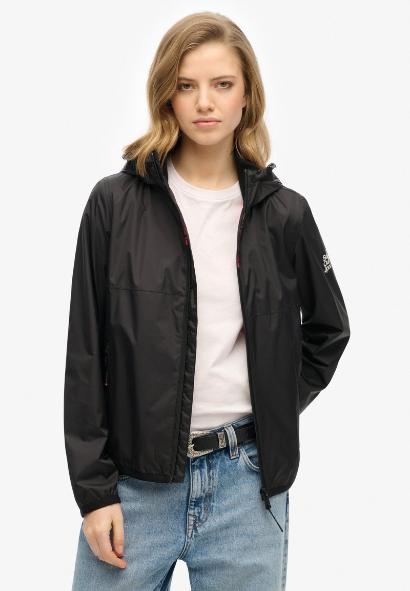 Veste de pluie légère noire avec capuche, fermeture éclair et deux poches latérales. Elle présente une texture lisse et un logo de marque subtile sur la manche.