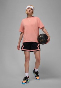 Rosa T-skjorte med Jordan-logo, svarte shorts med rosa detaljer, og fargerike treningssko. Holder en svart basketball med gull detaljer.