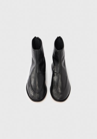 Stivali alla caviglia in pelle nera con una texture liscia, punta arrotondata e design minimalista. Caratterizzati da una cerniera laterale e dettagli di cucitura discreti.