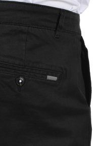 Schwarze Baumwollhosen mit einer geknöpften Tasche, die mit einem gestreiften Stoffakzent und dezenten Nähten entlang des Bundes und der Taschen versehen sind.