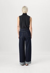 Top sin mangas gris con cuello alto, combinado con jeans de pierna ancha de denim oscuro, que presentan puños doblados y bolsillos traseros con parche.