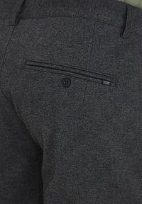 Pantalones gris oscuro confeccionados en tejido texturizado, con un bolsillo trasero que cuenta con un cierre de botón y una pequeña etiqueta de marca en el forro del bolsillo.