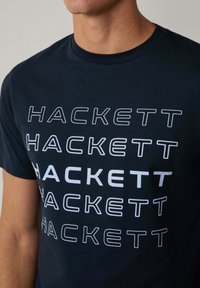 Námořnická modrá bavlněná tričko s více bílými logy "HACKETT" s různou tloušťkou a zarovnáním, kulatý výstřih, krátké rukávy.