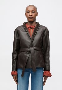 Καφέ δερμάτινο μπουφάν με στυλ περιτυλίγματος, αφράτο γιακά και ζώνη. Έχει κόκκινες λεπτομέρειες στους μανσέτες και υφή. Επίπεδο μπροστινό μέρος και μακριά μανίκια.