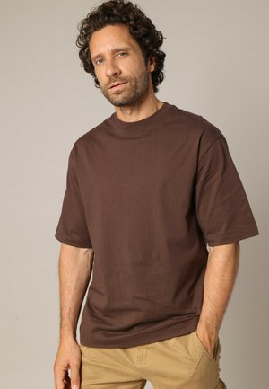 Homme aux cheveux bouclés portant un t-shirt marron à manches courtes et un pantalon beige, se tenant avec une main dans sa poche devant un fond uni.