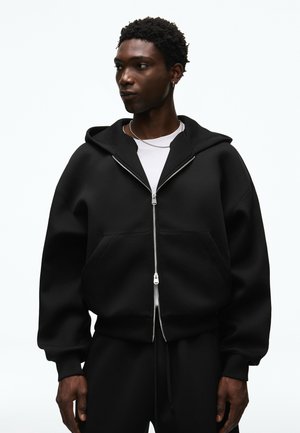 Giovane uomo che indossa una felpa nera oversize con zip e pantaloni neri, guarda di lato, su uno sfondo bianco semplice.
