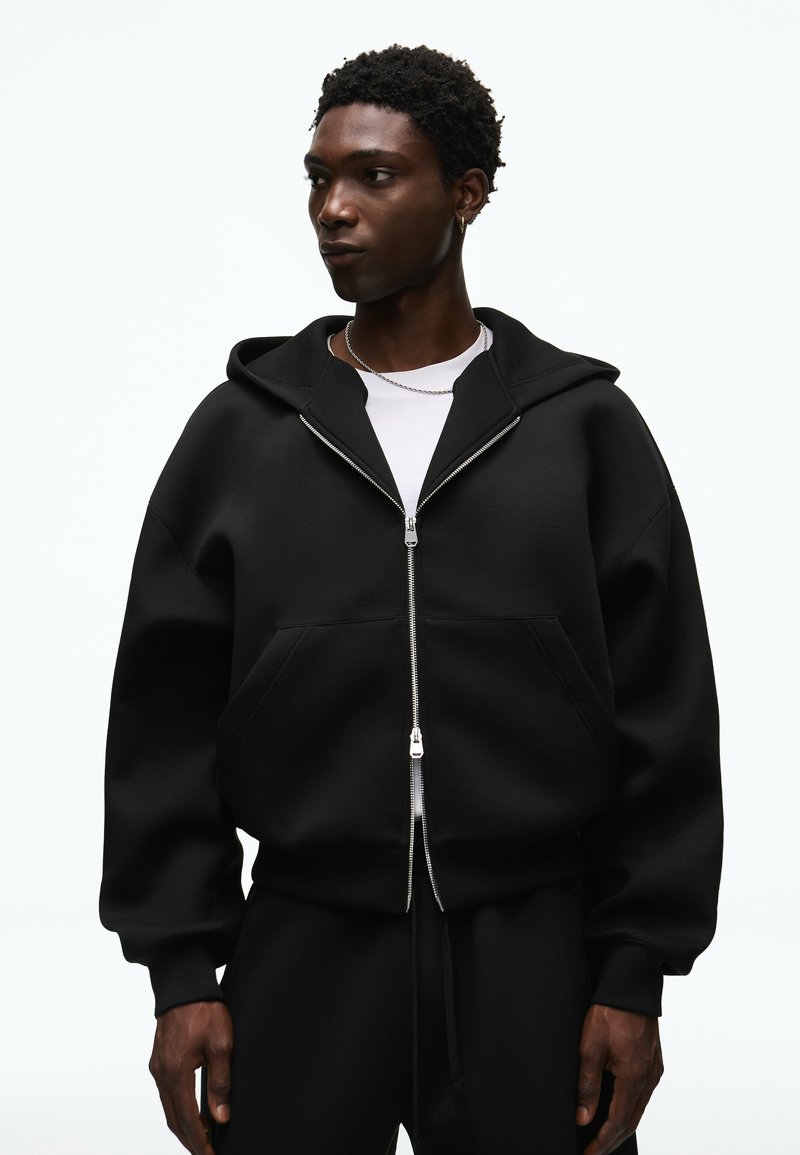 Jeune homme portant un sweat à capuche noir oversize zippé et pantalon noir, regardant sur le côté, sur un fond blanc uni.