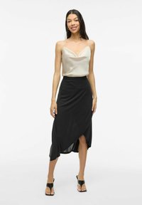 Beige satijnen top met dunne bandjes, gecombineerd met een zwarte wikkelrok met een asymmetrische zoom en een gladde textuur. Zwarte hoge sandalen.