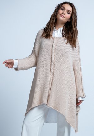 Beżowy sweter kardigan z dzianiny o teksturowanym wykończeniu, z luźnym, asymetrycznym krojem i szerokimi rękawami. Nosi się go na białą koszulę.