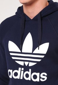 Námořnická modrá mikina s kapucí vyrobená z bavlněné směsi, s velkým bílým logem Adidas a třemi pruhy, s černou šňůrkou pro nastavení kapuce.