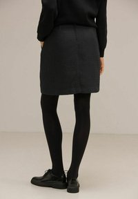 Svart texturerad kjol med en åtsittande design, med en söm bak. Bärs med svarta strumpbyxor och polerade skor. Neutral bakgrund.