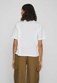 Cămașă albă din bumbac, cu mâneci scurte, decolteu rotund, croială lejeră și tiv drept, purtată cu pantaloni cargo maro, cu croială largă.