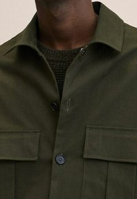 Camicia verde scuro con bottone frontale, due tasche sul petto e colletto a punta. Realizzata in un tessuto liscio di peso medio.