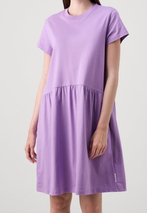 Vrouw draagt een losse, korte mouw, paarse jurk met een geplooide taille en een ronde halslijn tegen een eenvoudige lichte achtergrond.