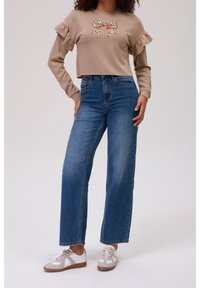 Suéter corto beige con mangas acampanadas y detalle de lazo de estampado de leopardo, combinado con jeans de mezclilla azul de tiro alto y zapatillas blancas.