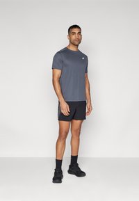 Graues Sport-T-Shirt mit kurzen Ärmeln, kombiniert mit schwarzen Shorts. Das Material scheint leicht zu sein und es hat ein kleines Logo auf der Brust.