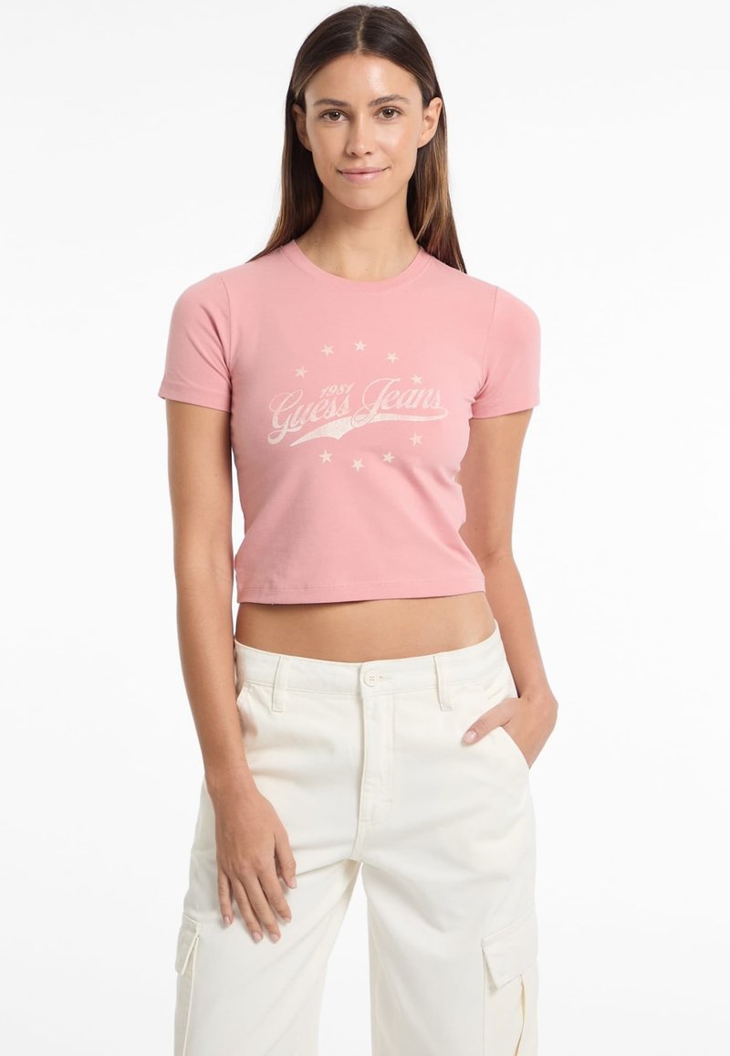Rožinė trumpa t-marškinė su trumpomis rankovėmis, kuriame pavaizduotas baltas "Guess Jeans" tekstas ir žvaigždžių raštas, pagaminta iš minkštos, tamprios medvilnės medžiagos.