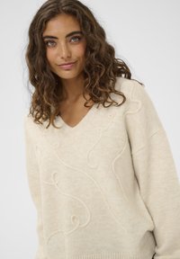 Krema krāsas džemperis ar V-veida kakla izgriezumu un mīkstu tekstūru. Priekšpusē ir spirālveida izšuvumu raksti un ribota apakšmalas apdare.