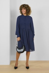 Abito blu marina a bottoni con tessuto testurizzato, vita arricciata e maniche lunghe. Il modello tiene in mano una borsa a mezzaluna nera, indossando tacchi a punta neri.