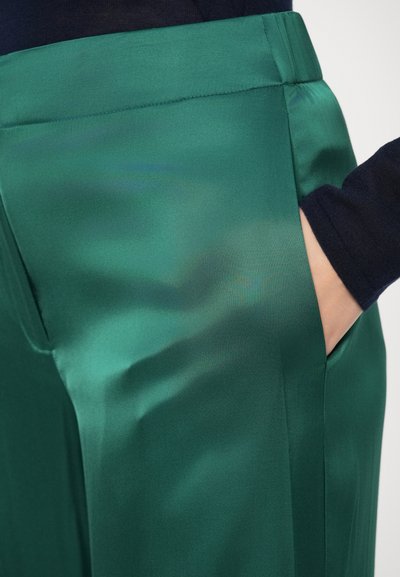Rohelised satiinsed püksid, millel on sile tekstuur, küljetasku ja lõigatud vöökoht. Kangas on pehme ja luksusliku läikega.