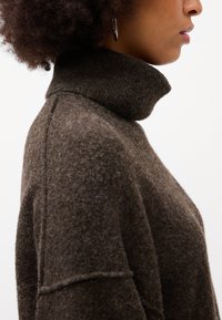 Maglione a collo alto in lana marrone, con una finitura testurizzata e dettagli di cucitura visibili, esposto su uno sfondo neutro.