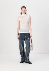 Hvit ermeløs topp med høy hals, kombinert med blå denimbukser med vertikale sølvglidelåsdetaljer. Modellen holder en lys grå håndveske.