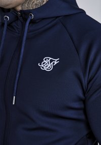 Námornícky zipsový mikina vyrobená z hladkej tkaniny, s bielym vyšitým logom, nastaviteľnými šnúrkami a striebornými kovovými akcentmi.