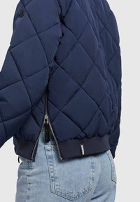Chaqueta de plumón navy con acolchado en diamante, bajo acanalado y cierre de cremallera. Presenta una cremallera negra y un detalle de etiqueta en la parte baja de la espalda.