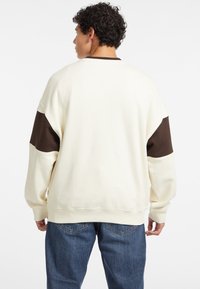 Sudadera de cuello redondo en color crema con acentos marrones en los hombros. Fabricada con material suave, presenta un ajuste relajado y puños acanalados.
