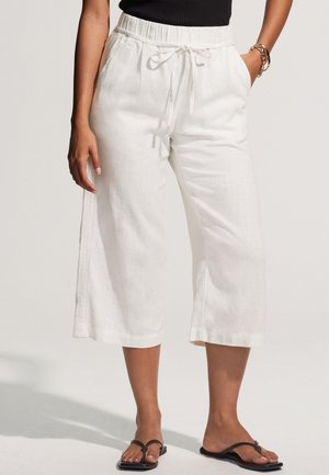 Nő magasderekú, széles szárú, fehér, rövidített nadrágot visel, megkötővel a derekán, fekete ujjatlan felsőt, arany karkötőt és fekete szandált.