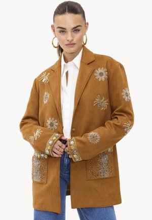 Femme portant une veste en daim brown avec des motifs de soleil et d'araignée en perles, accompagnée de boucles d'oreilles dorées en forme de créole, d'une chemise blanche et de jeans bleus.