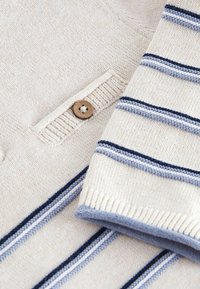 Pull en maille crème avec des rayures horizontales bleu marine et bleu clair, bouton en bois sur le col côtelé et poignets côtelés.